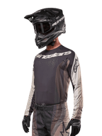 Maillot Alpinestars Techstar Pneuma 2024, maillot de motocross, sable foncé et gris fer, haut de performance à manches longues avec perforations au laser et logos blancs, porté avec un pantalon assorti et un casque noir, coupe athlétique pour le pilotage tout-terrain