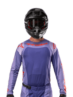 Maillot Alpinestars Techstar Ocuri 2024, violet et orange vif, porté par un pilote de motocross avec un casque et des lunettes noirs, doté de perforations découpées au laser sur la poitrine, d'une coupe athlétique de course et de logos rouges sur les épaules et le torse.