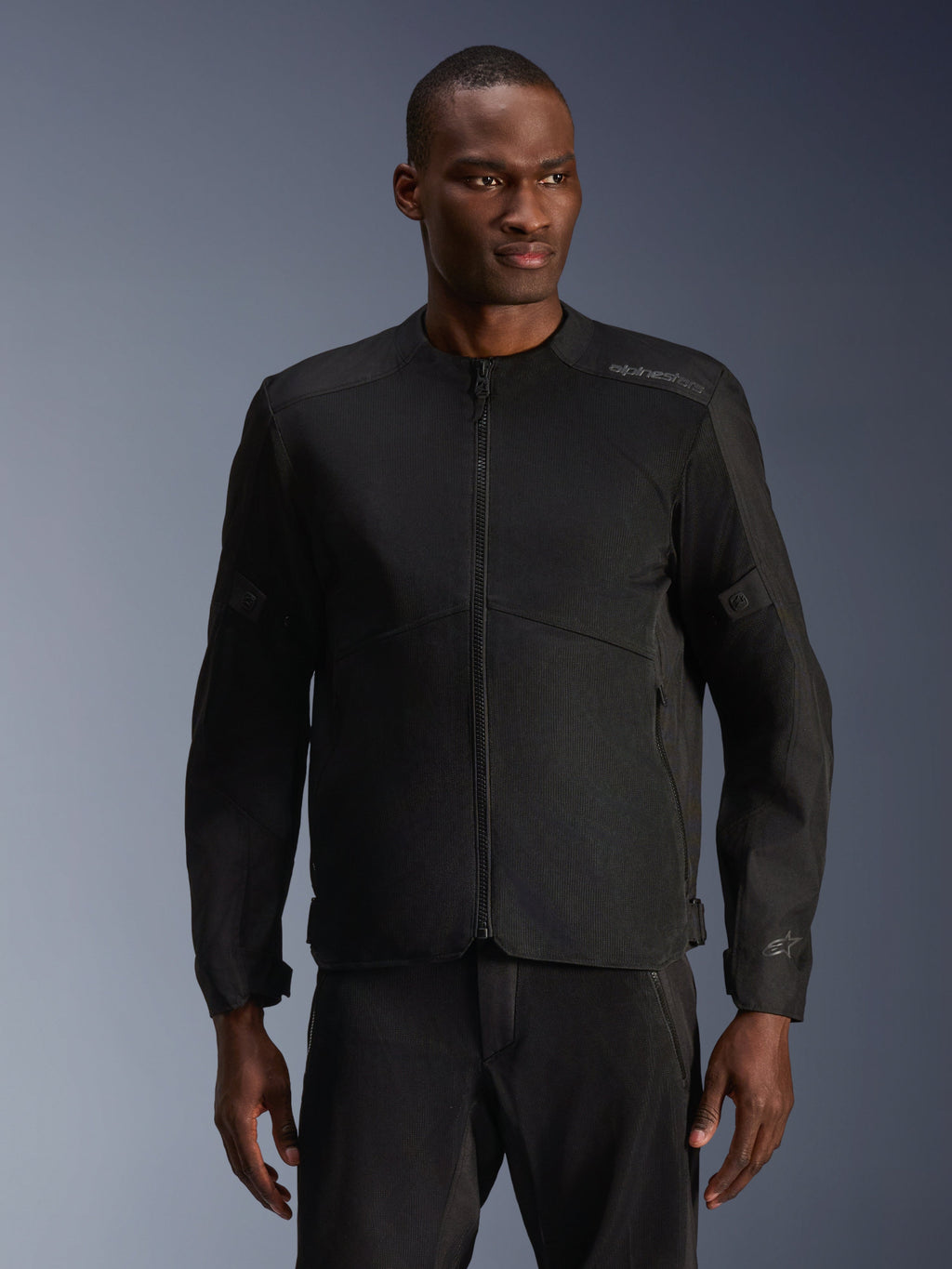 Mannequin homme portant la veste Alpinestars C-1 Air noire, une veste de moto contemporaine dotée de larges panneaux en maille technique et d'une fermeture éclair frontale intégrale, associée à un pantalon de moto noir assorti, esthétique urbaine et trajets quotidiens