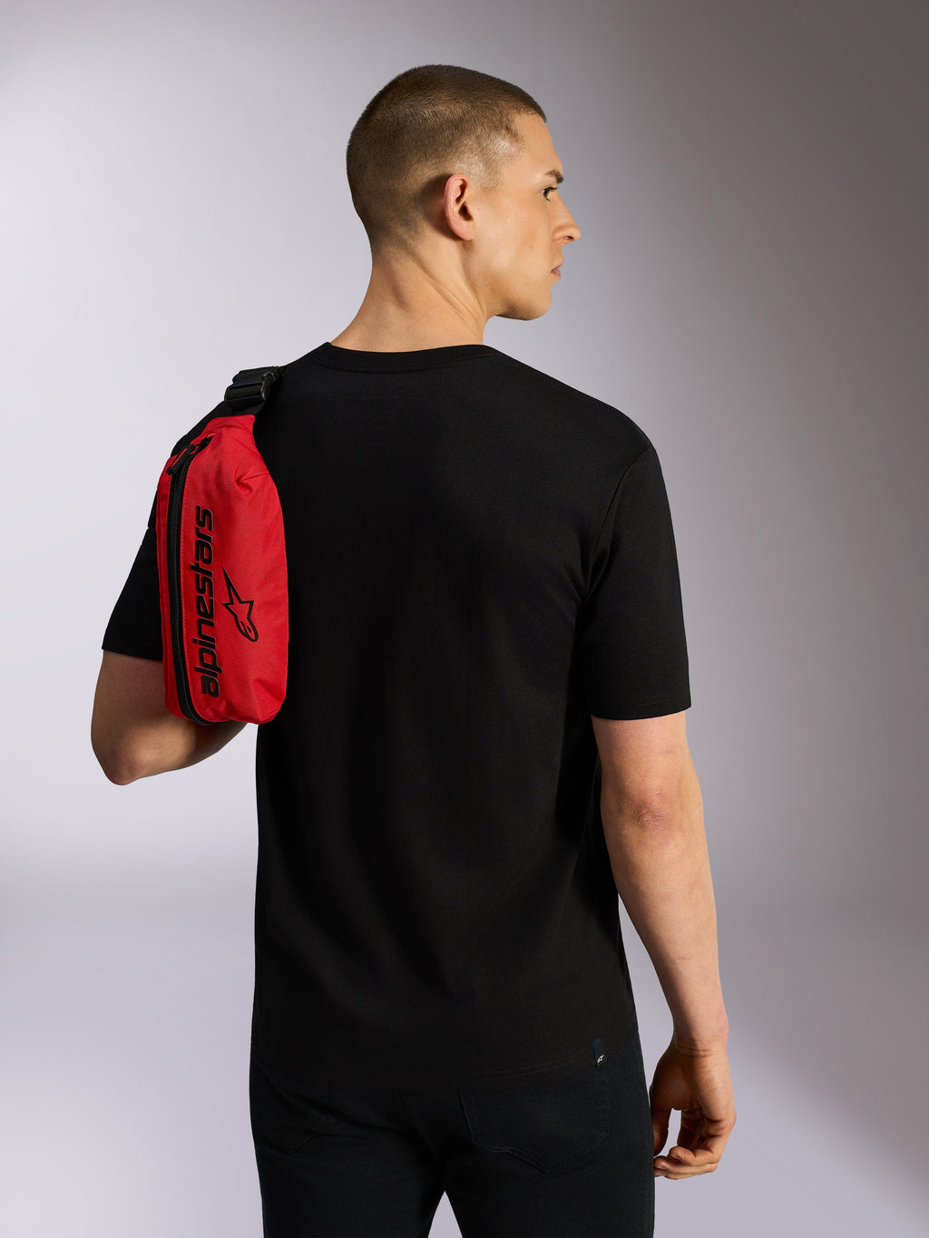 Sacoche de hanche Alpinestars Only Linear, rouge et noir, portée en bandoulière sur l'épaule par un mannequin en t-shirt noir, avec fermeture éclair verticale et logo Alpinestars noir, accessoire urbain durable.