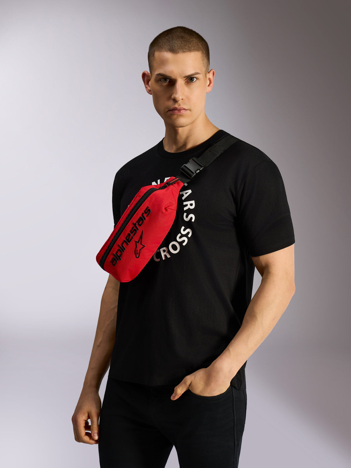 Sacoche de hanche Alpinestars Only Linear en rouge et noir, portée en bandoulière sur un t-shirt graphique noir, arborant un logo Alpinestars noir et une sangle réglable pour un look urbain décontracté.