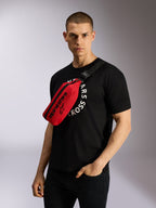 Sacoche de hanche Alpinestars Only Linear en rouge et noir, portée en bandoulière sur un t-shirt graphique noir, arborant un logo Alpinestars noir et une sangle réglable pour un look urbain décontracté.