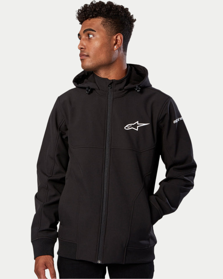 Alpinestars Primary Jacket, Black Softshell Jacket, male model wearing full-zip jacket with a hood, white Alpinestars logo on the chest and sleeve, ribbed cuffs and hem, technical motorsport-inspired streetwear